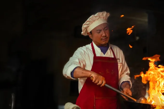 Chef de cozinha com avental vermelho em frente a um fogão, segurando uma panela. Cena do filme "Once Upon a Time in the Middle East".